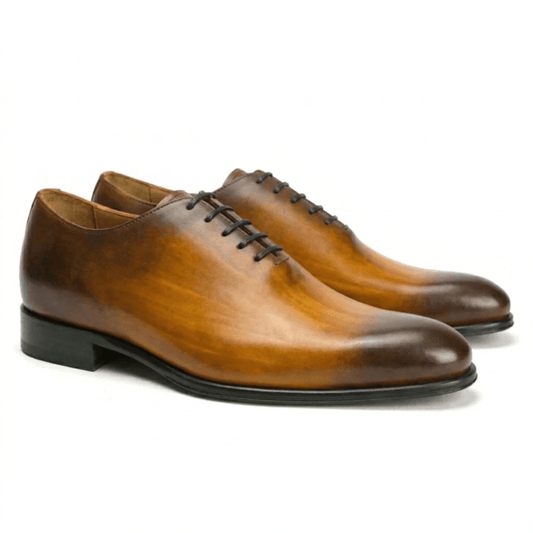 A pair of premium tan leather wholecut Oxford shoes featuring a seamless handcrafted design, a sophisticated dark burnished patina on the toe and heel, and elegant black lacing.