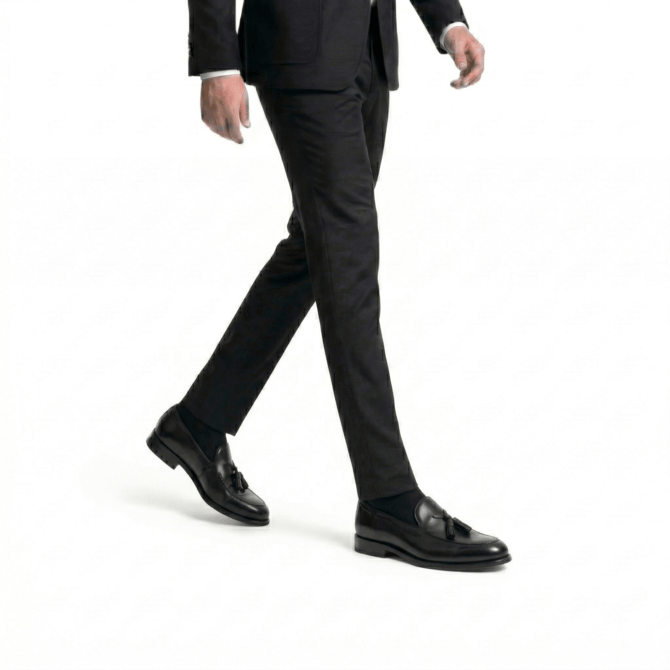 Lifestyle shot of black leather handcrafted tassel loafers paired with a tailored black suit, featuring a side-angle walking pose on a minimalist white background.