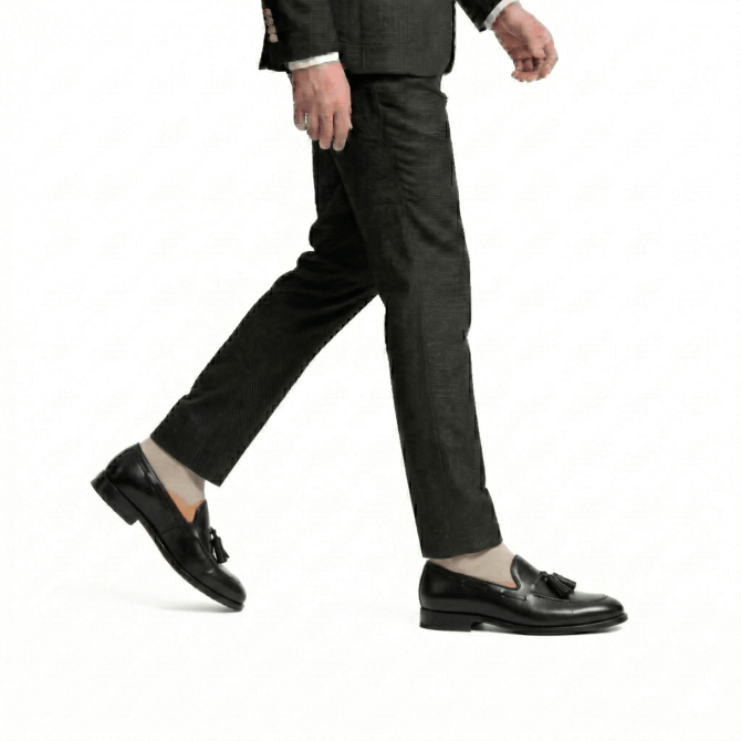 Side profile of a man walking in black leather tassel loafers and charcoal grey trousers, showcasing a minimalist professional style on a white background.