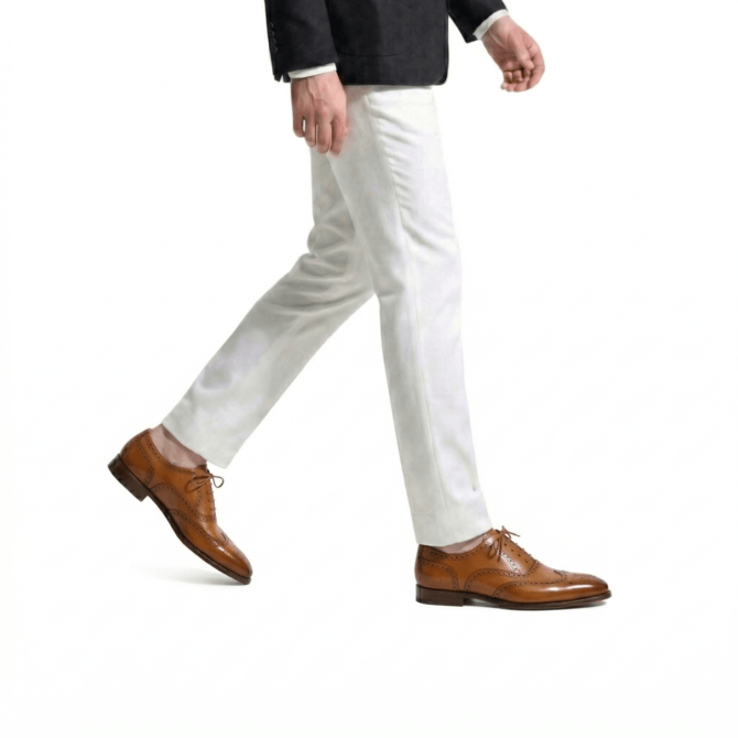 Side profile of a man walking in premium tan leather wingtip brogue shoes, paired with tailored white formal trousers and a black blazer.