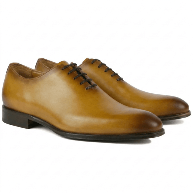 A pair of premium mustard-tan leather wholecut Oxford shoes featuring a seamless handcrafted design, a sophisticated burnished finish on the toe and heel, and elegant dark brown lacing.