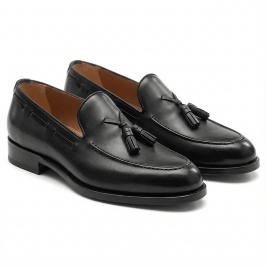 Three-quarter view of black leather tassel loafers for men, featuring a handcrafted apron toe and sleek tan leather lining on a minimalist white background.