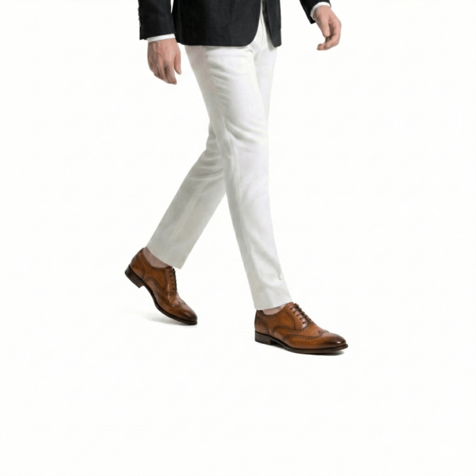 Side view of a man walking wearing a black textured blazer, slim-fit white formal trousers, and handcrafted tan leather wingtip brogue oxford shoes.