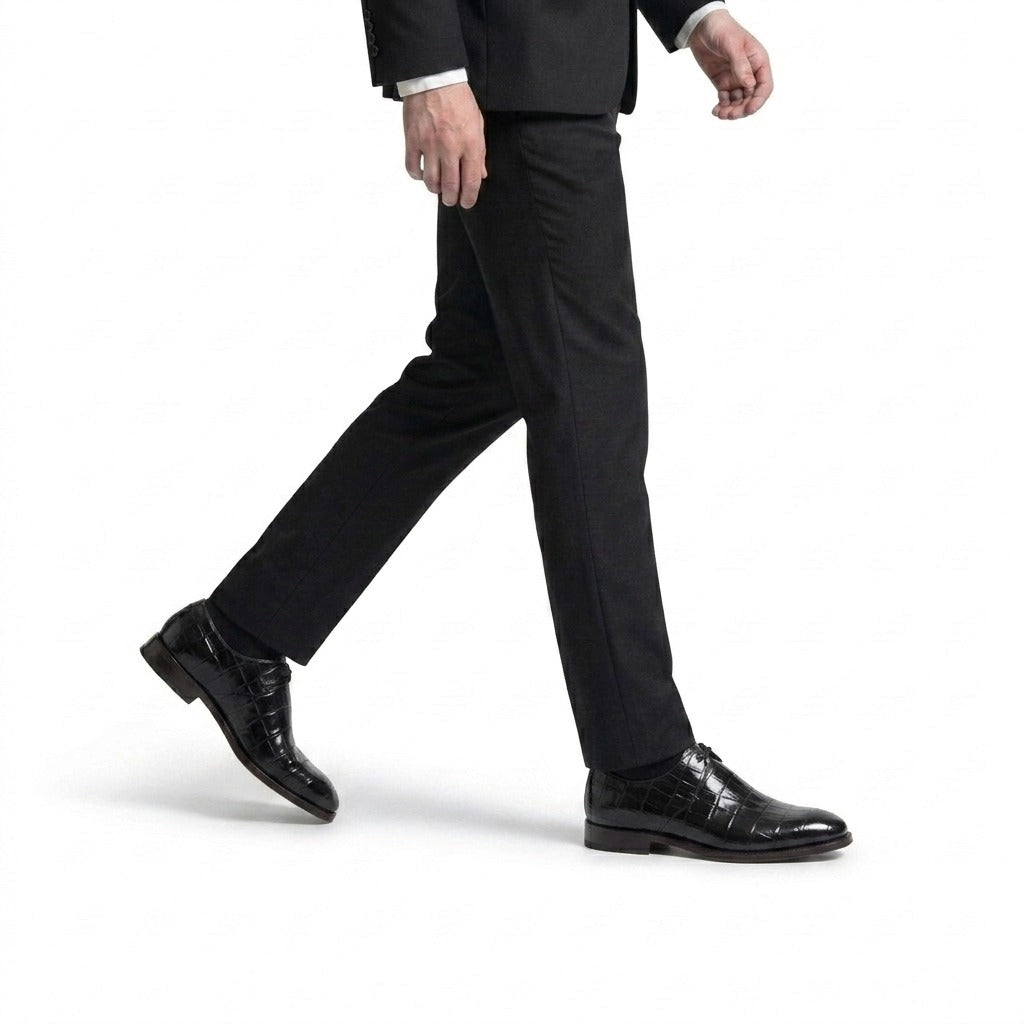 A side-angle view of a man walking in a tailored charcoal suit and luxury black crocodile-embossed leather derby shoes.