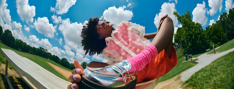 A fisheye wide-angle shot of a woman in a floral blouse and pink socks sitting on park bleachers, showcasing shiny silver metallic platform loafers against a bright blue sky.