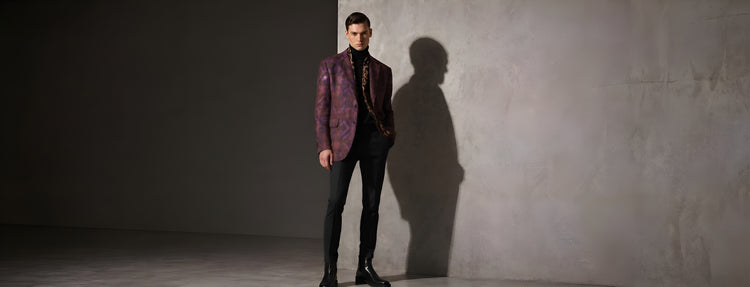 A male model wearing a luxury purple patterned blazer, black turtleneck, and sleek black leather Chelsea boots in a high-fashion studio setting.