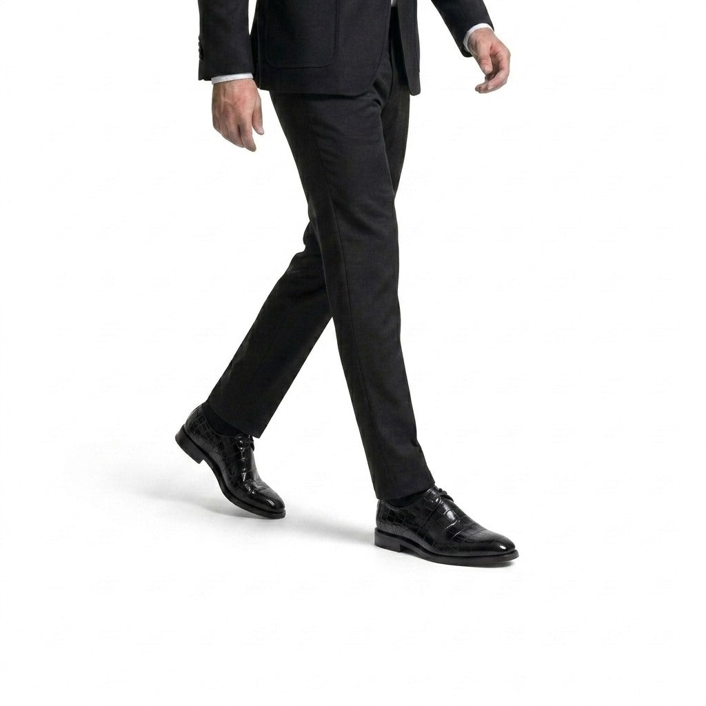 Side view of a man walking in black crocodile-embossed leather dress shoes and a tailored charcoal grey suit.
