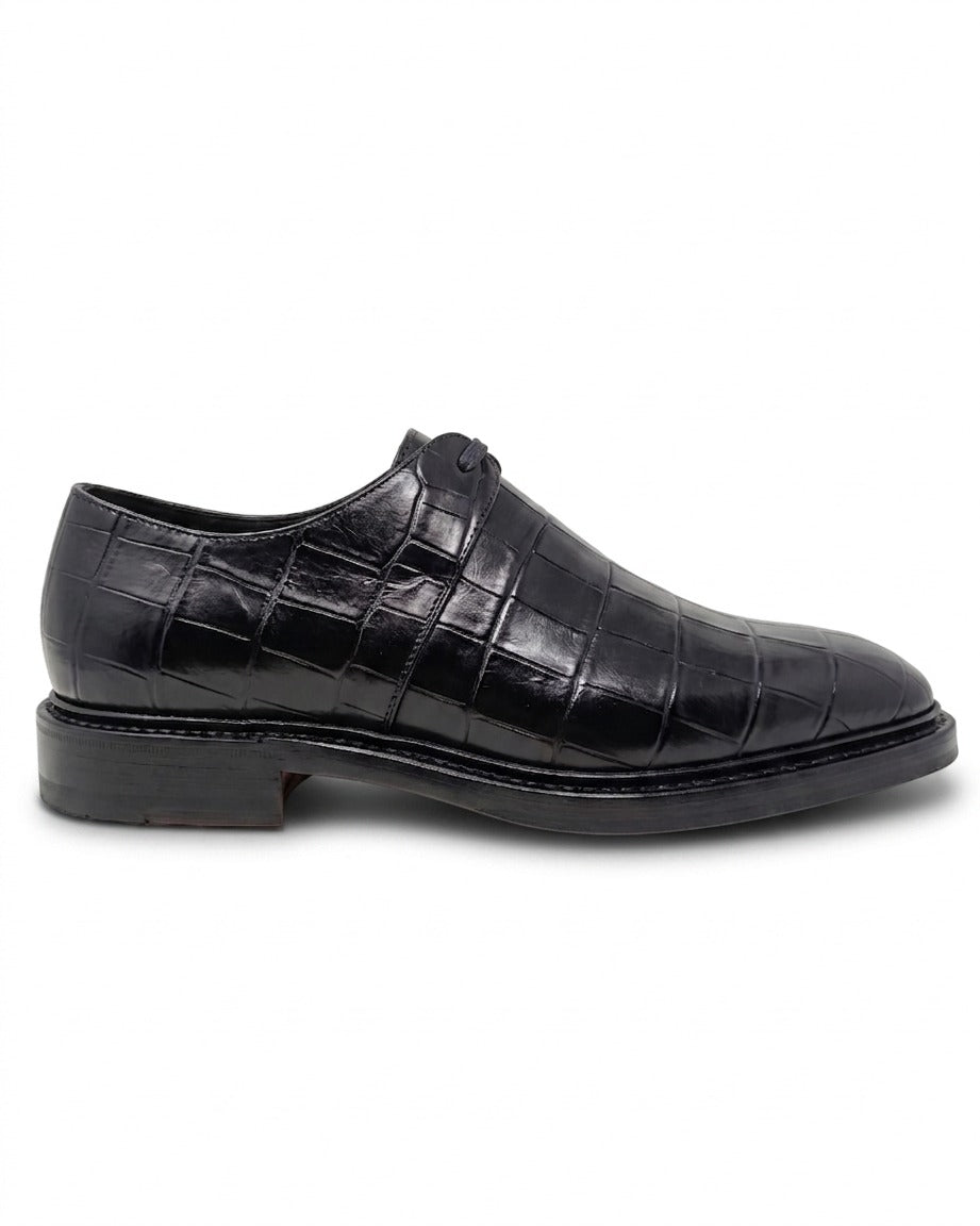 Side profile view of a black crocodile-embossed leather dress shoe featuring a low heel and stacked leather sole.